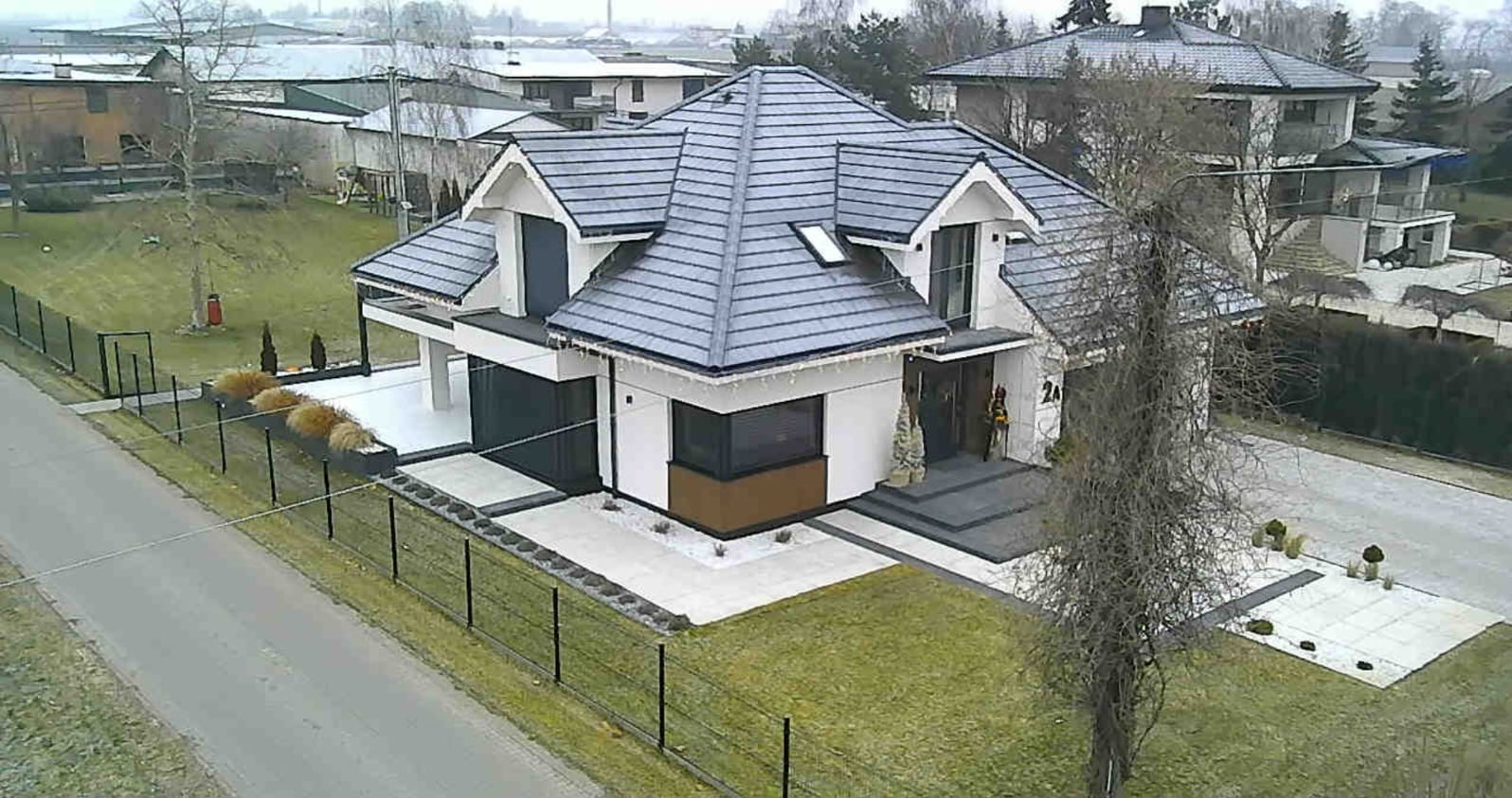 Nowoczesny dom jednorodzinny z grafitową dachówką, białymi ścianami i elementami w kolorze drewna. Widok z góry, ogrodzenie z czarnych prętów.