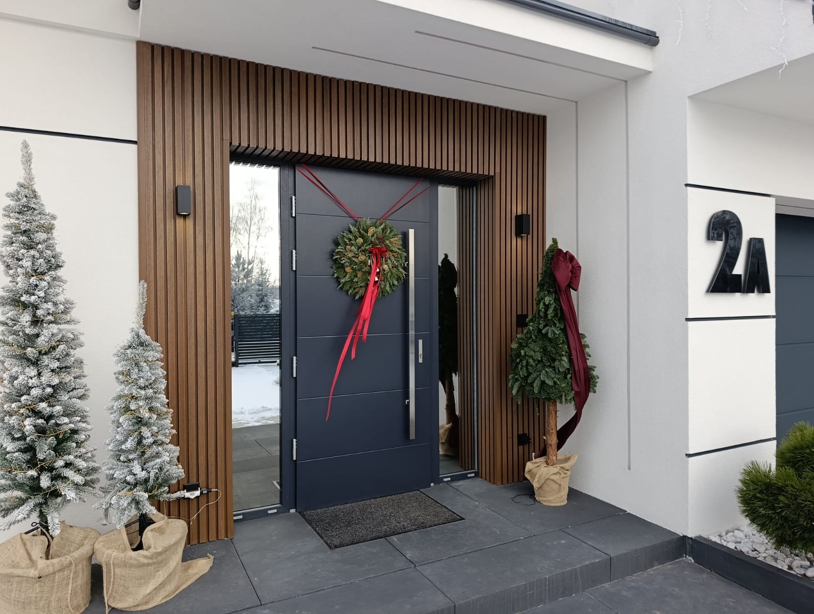 Nowoczesne wejście do domu z drewnianą okładziną, szarą stolarką i świątecznym wieńcem. Dwie choinki w jutowych workach dodają uroku.