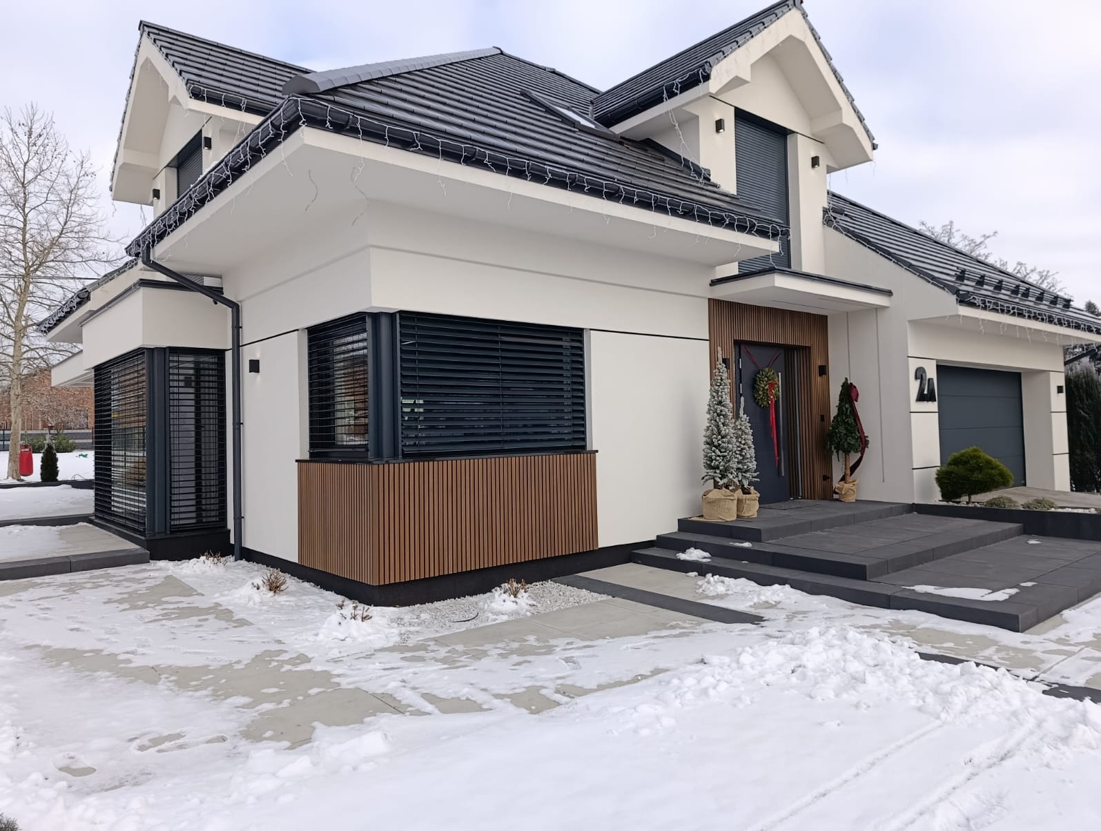 Nowoczesny dom jednorodzinny z białymi ścianami, drewnianą okładziną i szarym dachem w zimowej scenerii. Dekoracje świąteczne przy wejściu.