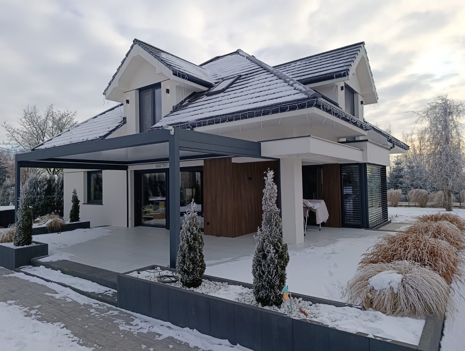 Nowoczesny dom jednorodzinny z pergolą w zimowej scenerii. Śnieg na dachu i roślinach, minimalistyczny design, drewniane akcenty elewacji.
