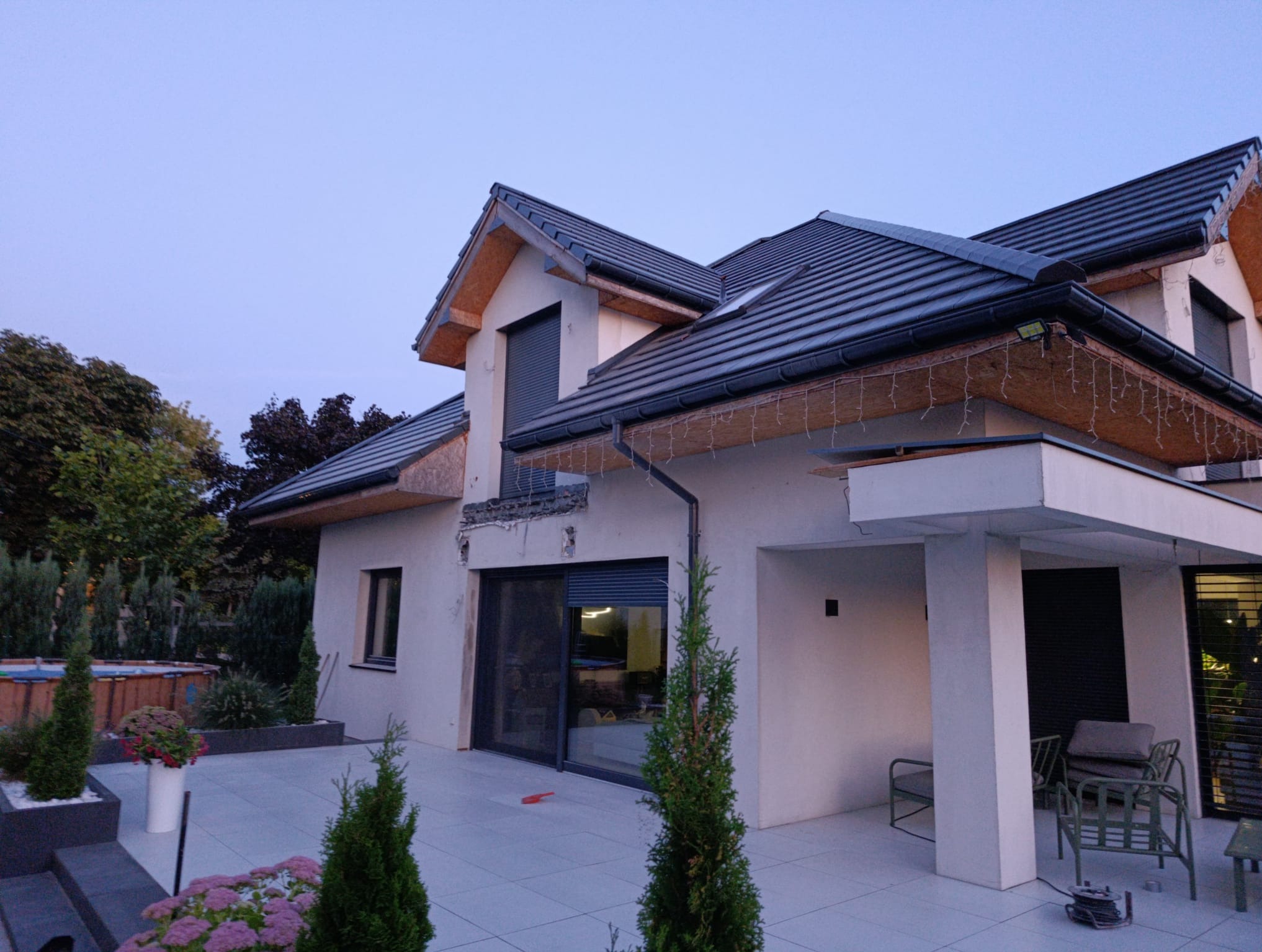 Nowoczesny dom jednorodzinny z jasną elewacją i ciemną dachówką, widok z zewnątrz na taras z roślinami i meblami ogrodowymi, oświetlenie zewnętrzne.