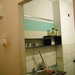 Waldemar Biela - Wnętrze kuchni w trakcie prac: widoczna niezabezpieczona ściana nad zlewem, częściowo pomalowana na zielono, białe płytki na ścianie, półki z przyprawami, widoczne ślady prac na ścianie po lewej...