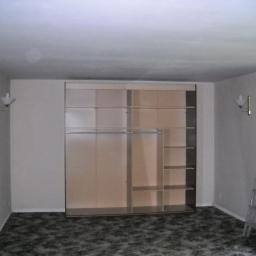 Waldemar Biela - Pokój z wbudowaną szafą z przesuwnymi drzwiami, widoczne półki i drążki na ubrania, drabina oparta o ścianę, wzorzysta wykładzina podłogowa, lampki ścienne.