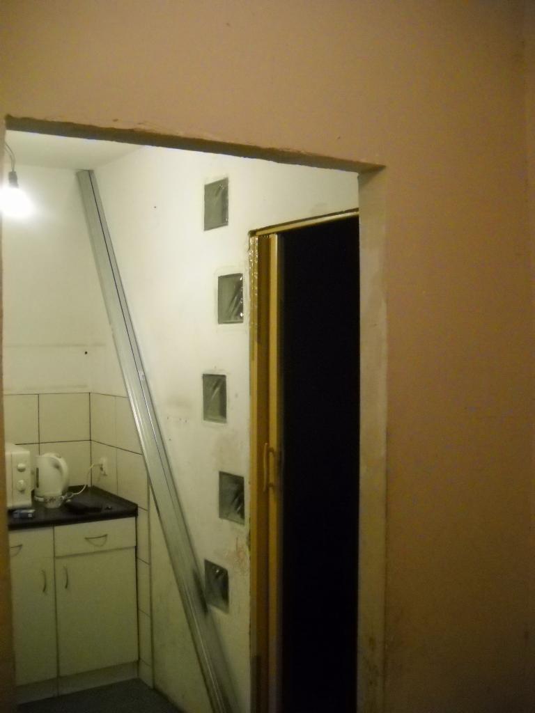 Wnętrze w trakcie remontu: widoczna kuchnia z białymi płytkami i szafkami, częściowo zasłonięta metalowym profilem, ściana z małymi okienkami i fragmentem drzwi, całość w surowym stanie przed...