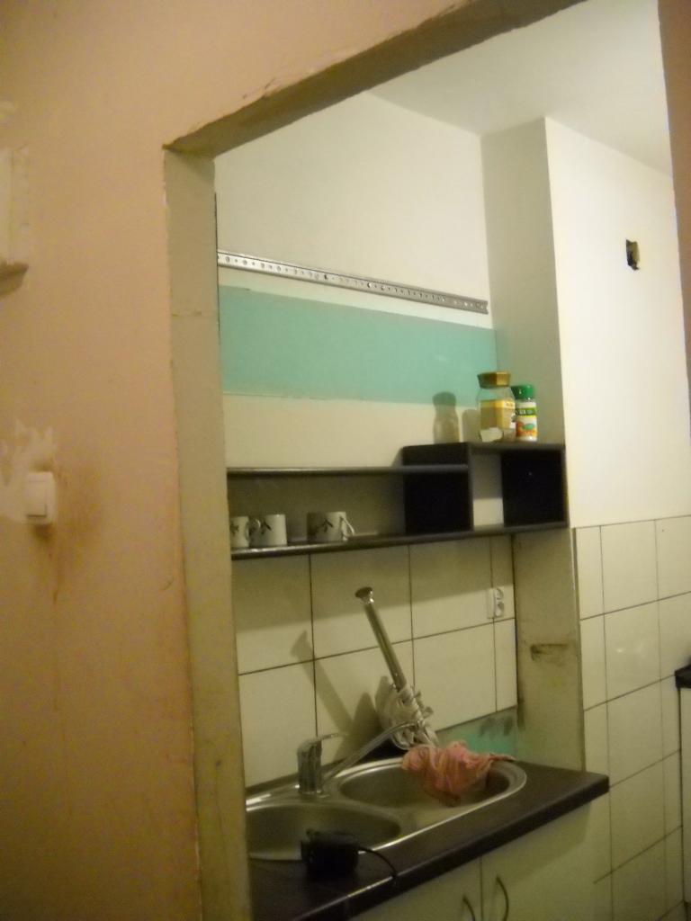 Wnętrze kuchni w trakcie prac: widoczna niezabezpieczona ściana nad zlewem, częściowo pomalowana na zielono, białe płytki na ścianie, półki z przyprawami, widoczne ślady prac na ścianie po lewej...