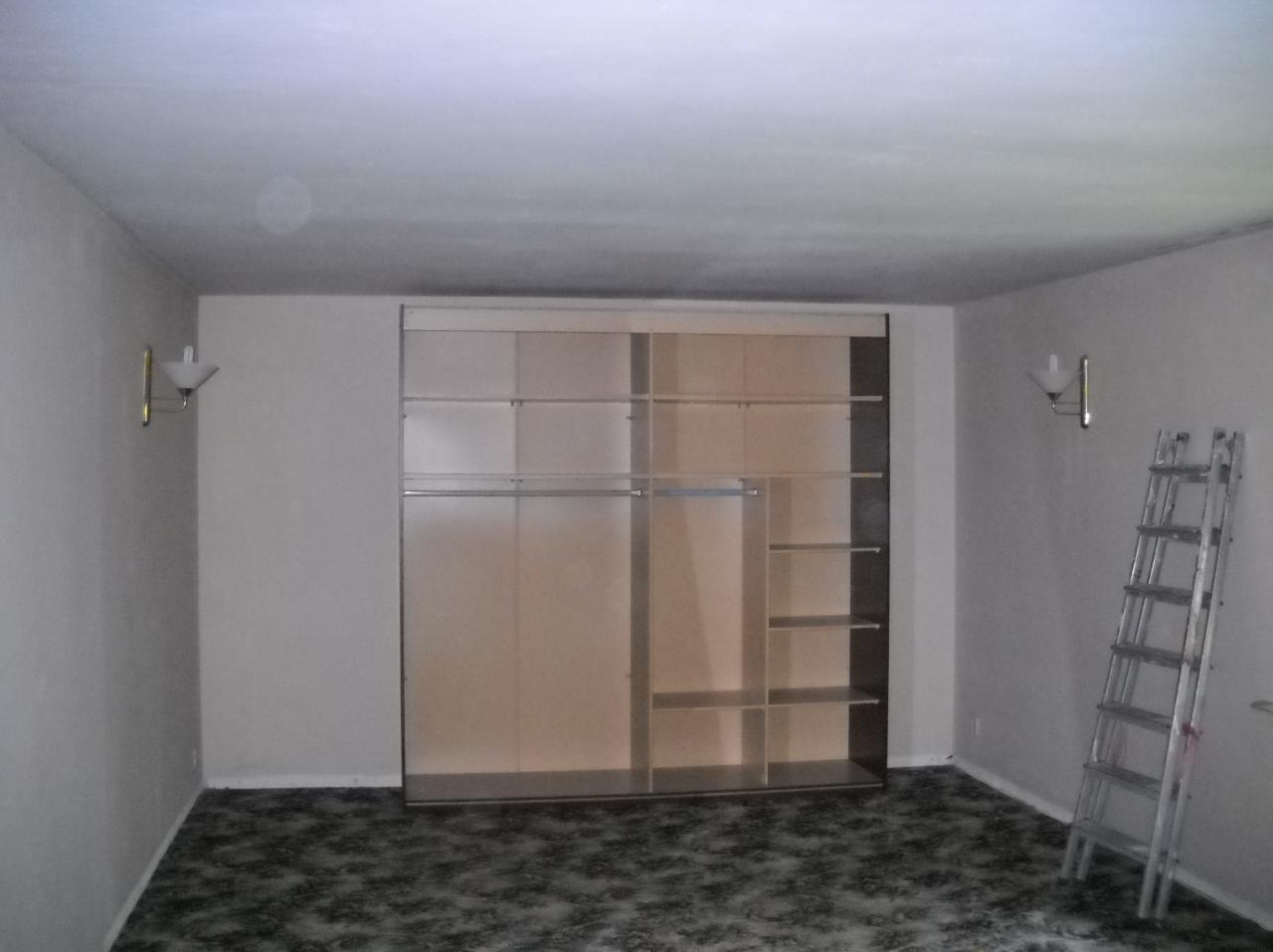 Pokój z wbudowaną szafą z przesuwnymi drzwiami, widoczne półki i drążki na ubrania, drabina oparta o ścianę, wzorzysta wykładzina podłogowa, lampki ścienne.