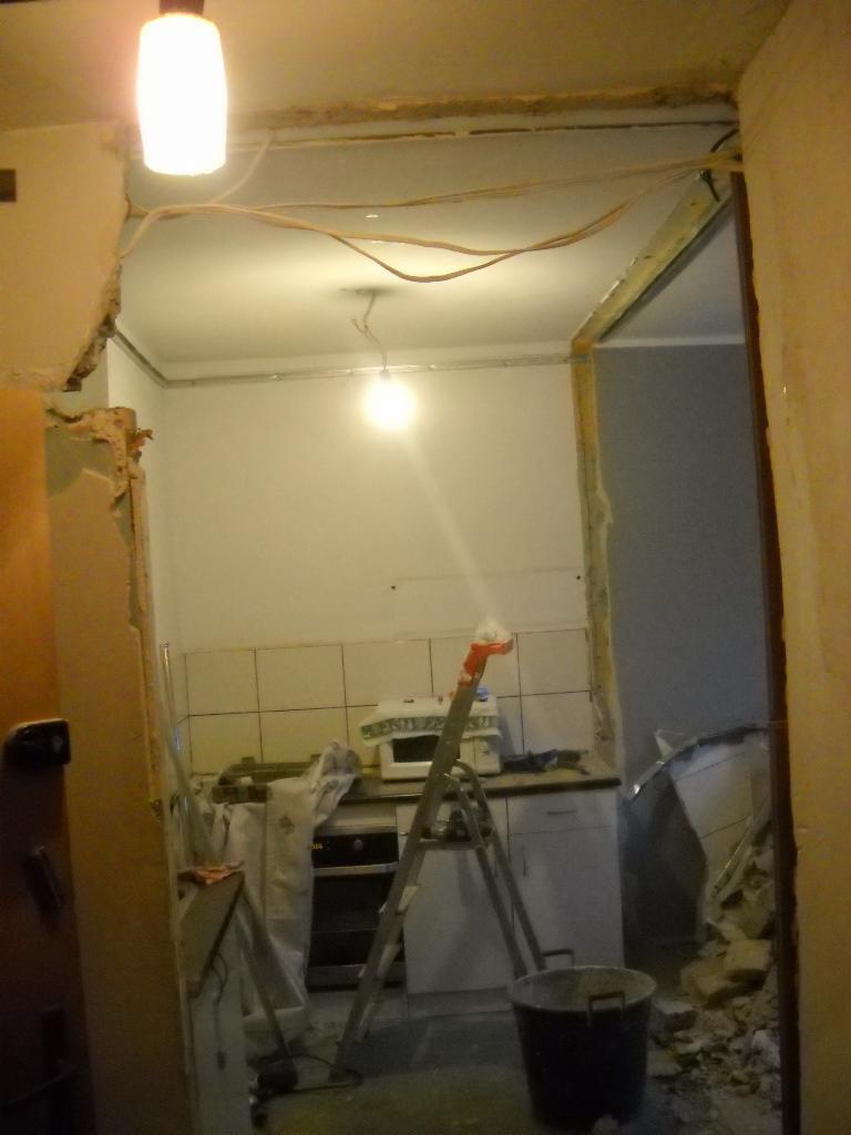 Demontaż w kuchni: widok na usunięte płytki, odsłonięte przewody elektryczne, drabina oparta o blat, wiadro na podłodze i fragmenty gruzu.
