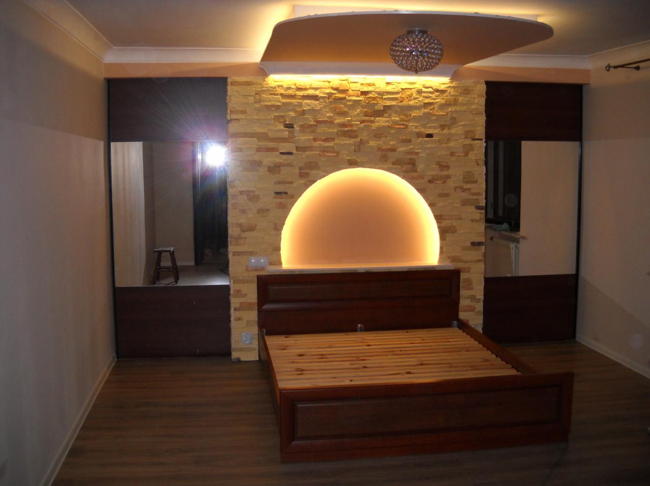 Sypialnia z łóżkiem z ciemnego drewna, ściana z dekoracyjnej cegły podświetlona półkolistym światłem, oraz szafy z lustrzanymi drzwiami po bokach.