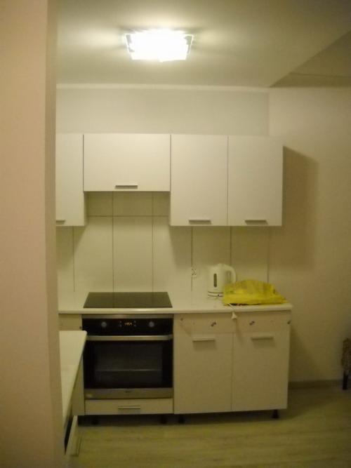 Mała kuchnia z białymi szafkami wiszącymi i stojącymi, czarną płytą indukcyjną i piekarnikiem, oraz białym czajnikiem elektrycznym i żółtą torbą na blacie.