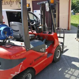 Czerwony wózek widłowy Suchedniów z butlą gazową, zaparkowany na kostce brukowej. Widoczny fragment białego busa i budynek w tle.