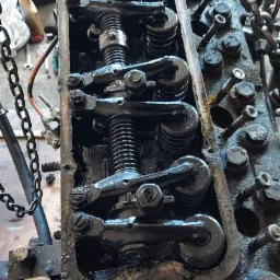 Głowica silnika wózka widłowego, widoczne sprężyny zaworowe i dźwigienki, podwieszona na łańcuchu. Detale mechaniczne, smar i ślady użytkowania.