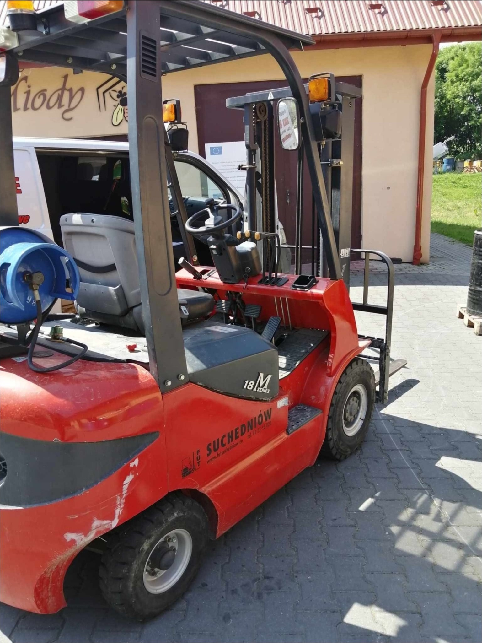 Czerwony wózek widłowy Suchedniów z butlą gazową, zaparkowany na kostce brukowej. Widoczny fragment białego busa i budynek w tle.