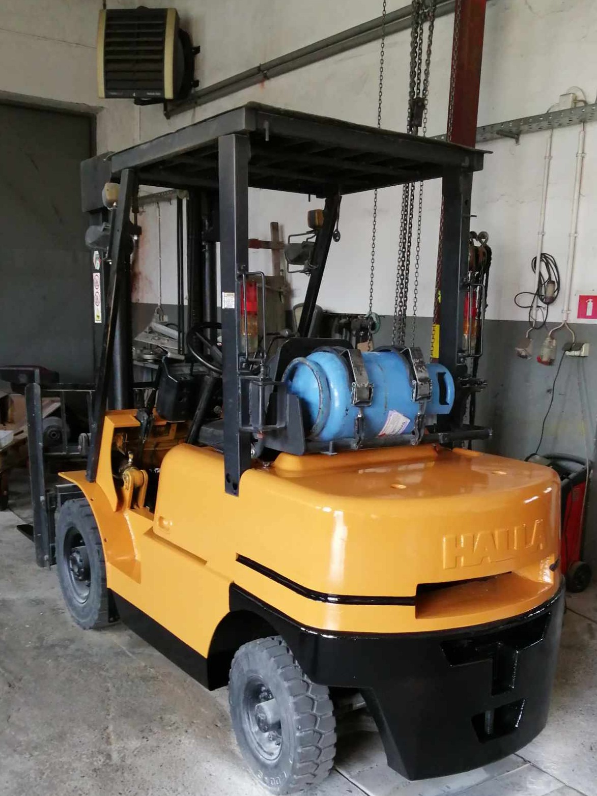 Żółto-czarny wózek widłowy z niebieską butlą gazową w warsztacie. Widoczne łańcuchy i narzędzia. Model HAILA.