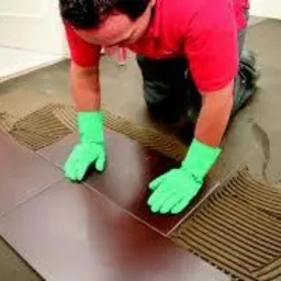 Mężczyzna w zielonych rękawicach układa brązowe płytki na podłodze pokrytej klejem. Widoczne rowki w kleju ułożone specjalną pacą. Częściowo ułożona podłoga w pomieszczeniu.