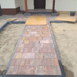 Układanie ścieżki z kostki brukowej w odcieniach brązu i czerwieni, obrzeża z szarego betonu, widoczne płyty ochronne i piasek, etap prac przed fugowaniem.