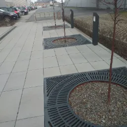 Miejsce zasadzenia drzew z ażurowymi osłonami i szarym żwirem na tle chodnika z jasnoszarych płyt i budynków przemysłowych w pochmurny dzień.