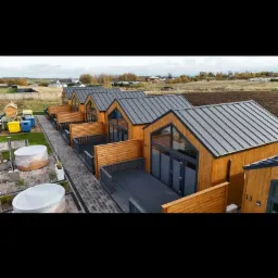 Kompleks kilku identycznych, drewnianych domków letniskowych z szarymi dachami i tarasami, otoczonych zielenią i z widocznymi jacuzzi na zewnątrz.