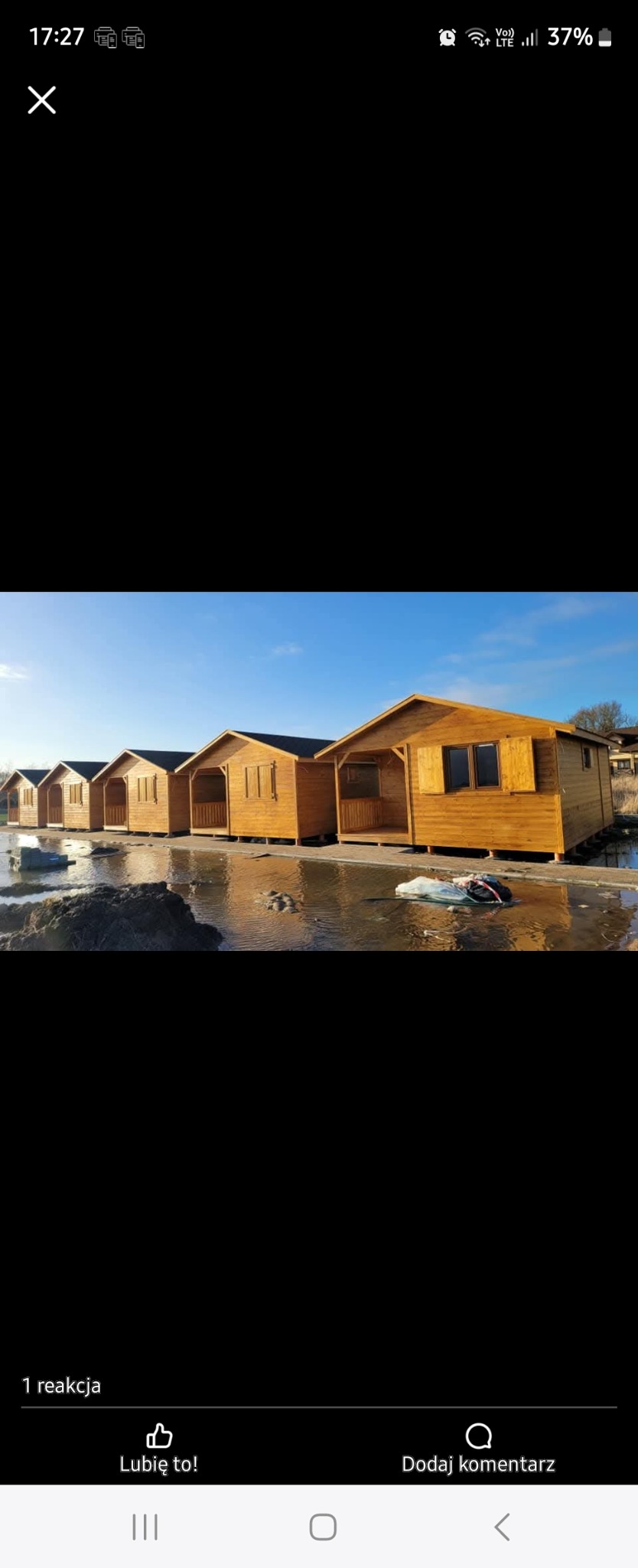 Szereg identycznych, surowych, drewnianych domków letniskowych z czarnymi dachami ustawionych w rzędzie na podmokłym terenie, odbijających się w wodzie.