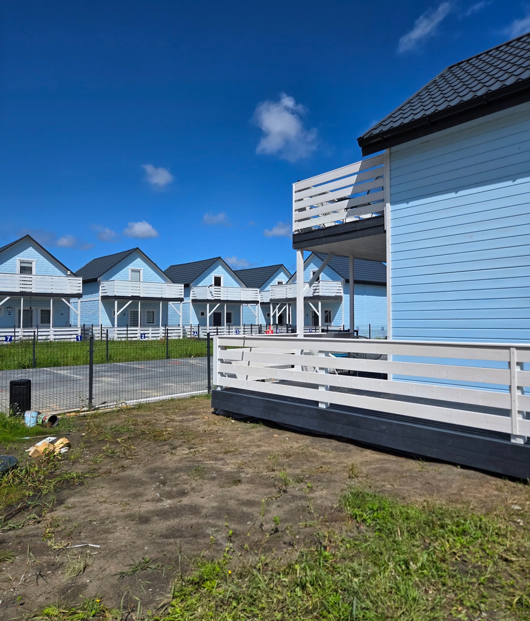 Kompleks kilku identycznych, parterowych domków letniskowych z balkonami, pomalowanych na jasnoniebiesko z białymi balustradami, widocznych z zewnątrz, na tle błękitnego nieba z chmurami.