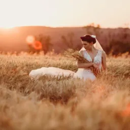 Panna młoda w białej sukni z bukietem kłosów pszenicy siedzi na polu zboża w promieniach zachodzącego słońca.