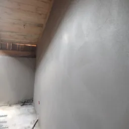 Świeżo otynkowana ściana z widoczną drewnianą konstrukcją sufitu poddasza i wystającą instalacją elektryczną w dolnej części ściany