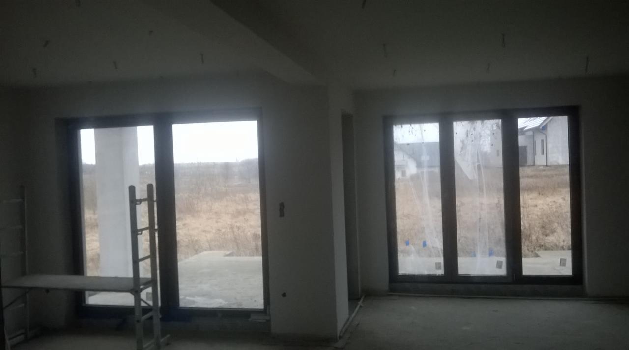 Wnętrze w stanie deweloperskim z dużymi oknami, widoczny aluminiowy podest malarski i surowy tynk na ścianach.