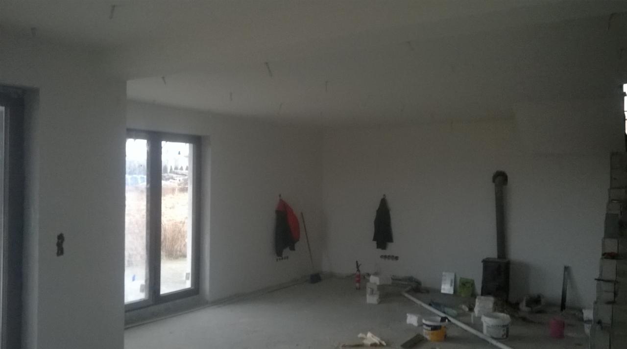 Wnętrze w trakcie prac wykończeniowych: ściany po tynkowaniu, widoczne narzędzia, wiadro z farbą, oraz ubrania robocze wiszące na ścianie.