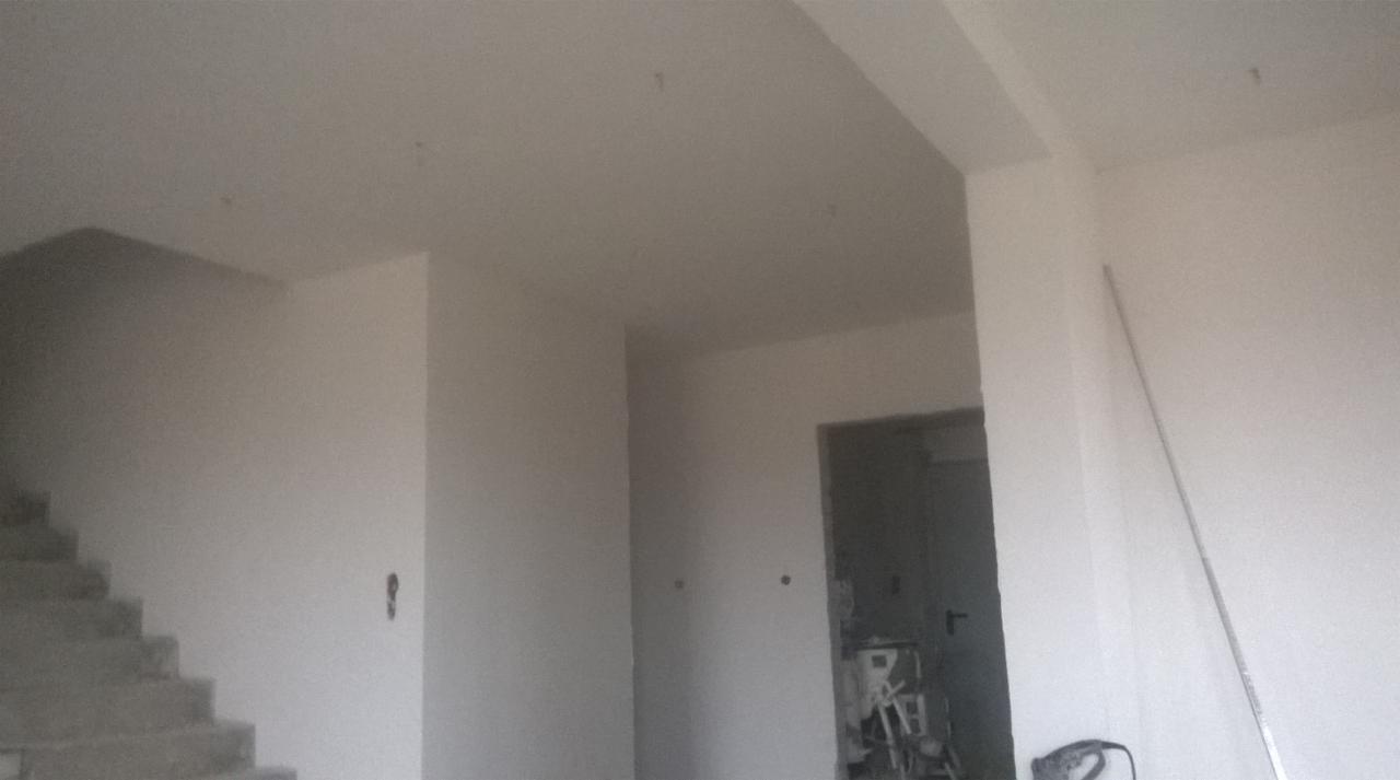 Wnętrze w trakcie prac wykończeniowych: świeżo otynkowane ściany, widoczne schody w surowym stanie, narzędzia malarskie w tle przy niezabezpieczonym otworze drzwiowym.