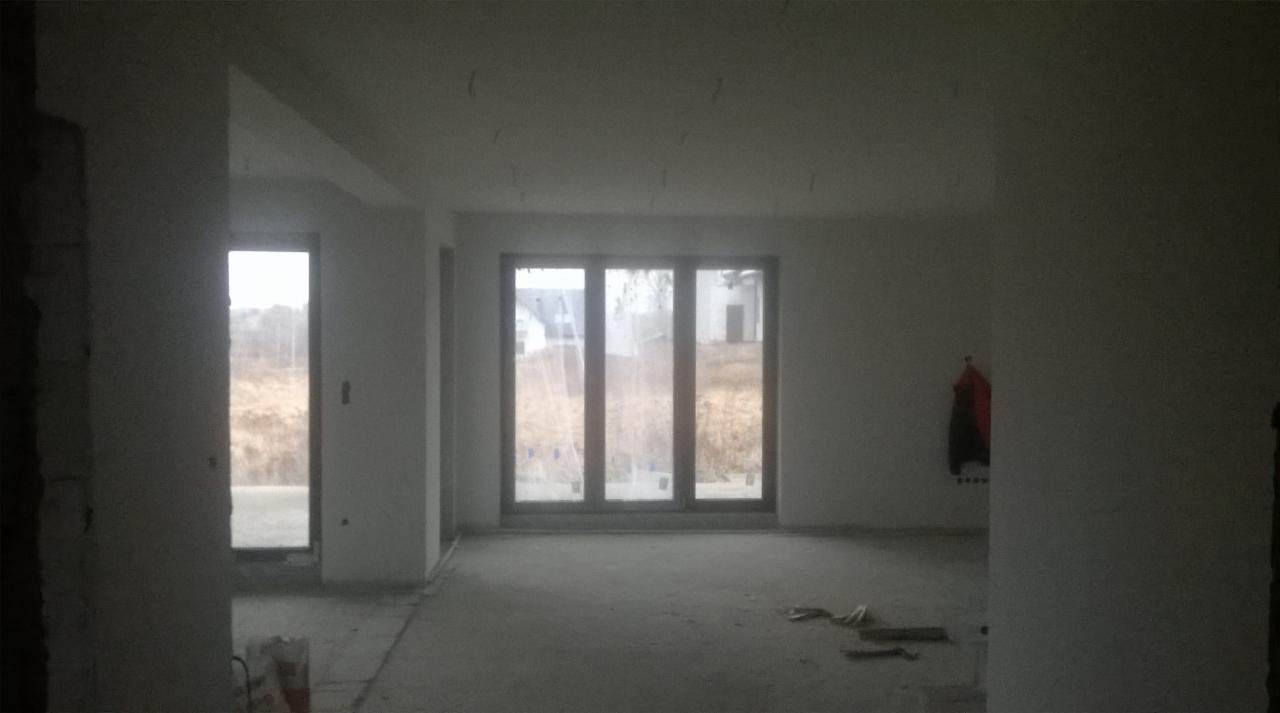 Wnętrze w stanie deweloperskim z dużym oknem balkonowym, ściany przygotowane do malowania, widoczne ślady po pracach wykończeniowych na podłodze, w tle widok na zewnątrz.