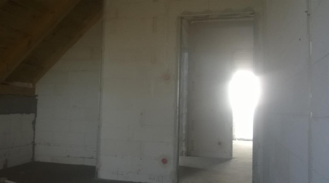 Wnętrze surowego poddasza ze ścianami z betonu komórkowego, widoczne drewniane elementy konstrukcyjne dachu, przygotowane do dalszych prac wykończeniowych, z intensywnym światłem wpadającym przez...