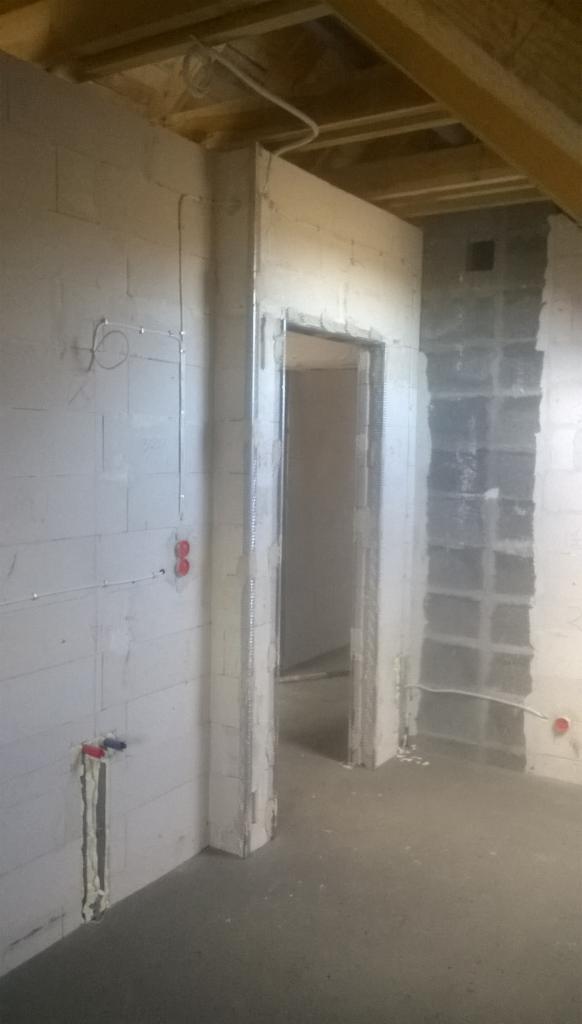 Wnętrze w stanie surowym: ściany z betonu komórkowego z widoczną instalacją elektryczną, metalowa ościeżnica drzwiowa przed montażem, sufit z drewnianych belek.