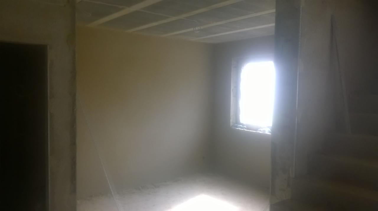 Świeżo otynkowane ściany w surowym wnętrzu, widoczne schody i okno z intensywnym światłem dziennym, sufit z widoczną konstrukcją nośną.