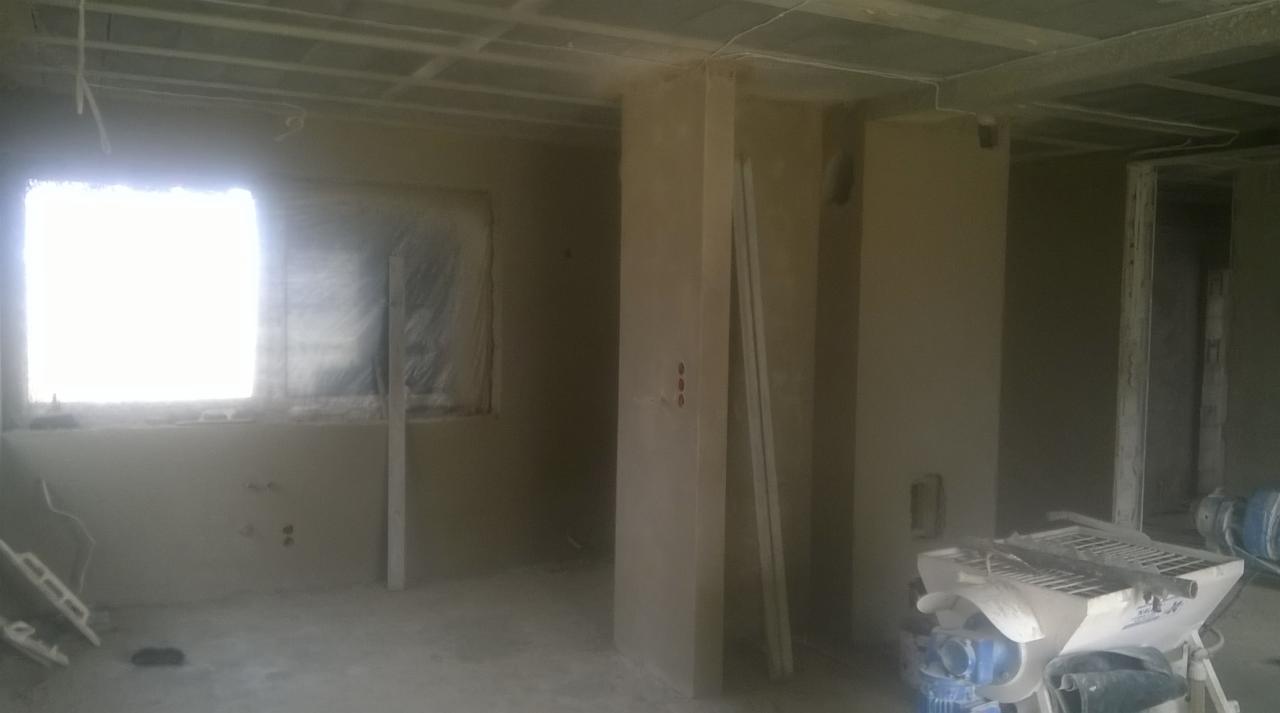 Wnętrze w trakcie prac wykończeniowych: ściany pokryte świeżym tynkiem, okno zabezpieczone folią, widoczna maszyna tynkarska i profile przyścienne.