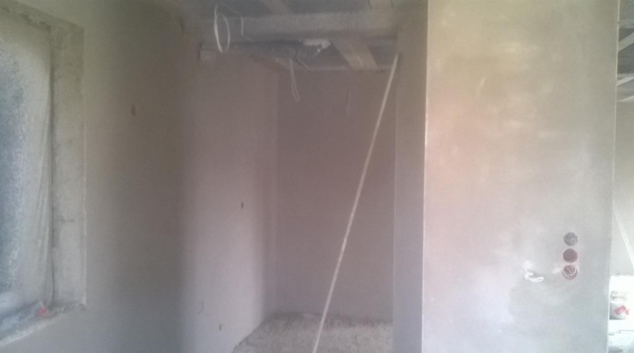 Wnętrze w trakcie prac wykończeniowych, świeżo otynkowane ściany z widocznymi puszkami elektrycznymi, sufit z widoczną konstrukcją nośną i przewodami.