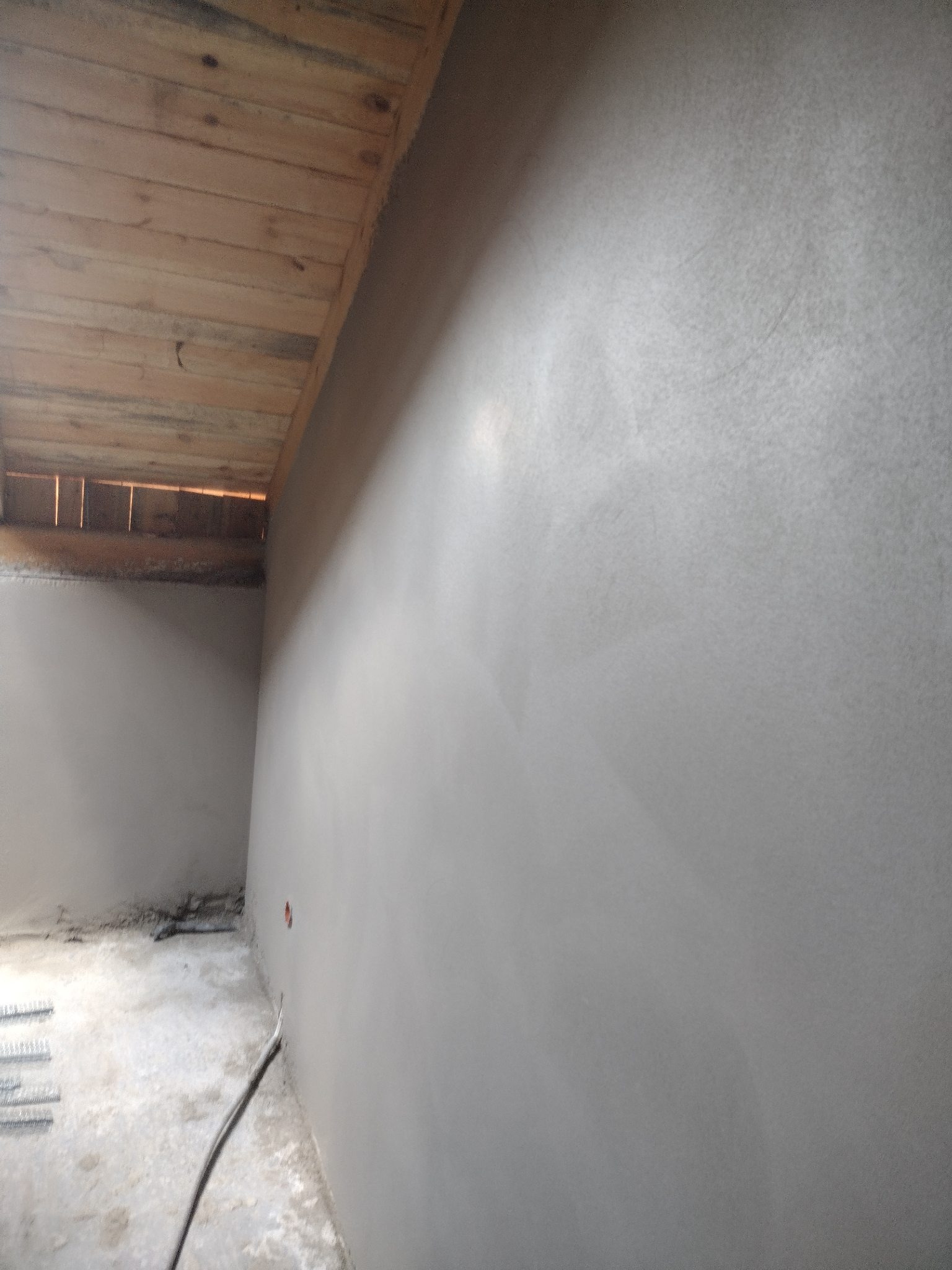 Świeżo otynkowana ściana z widoczną drewnianą konstrukcją sufitu poddasza i wystającą instalacją elektryczną w dolnej części ściany