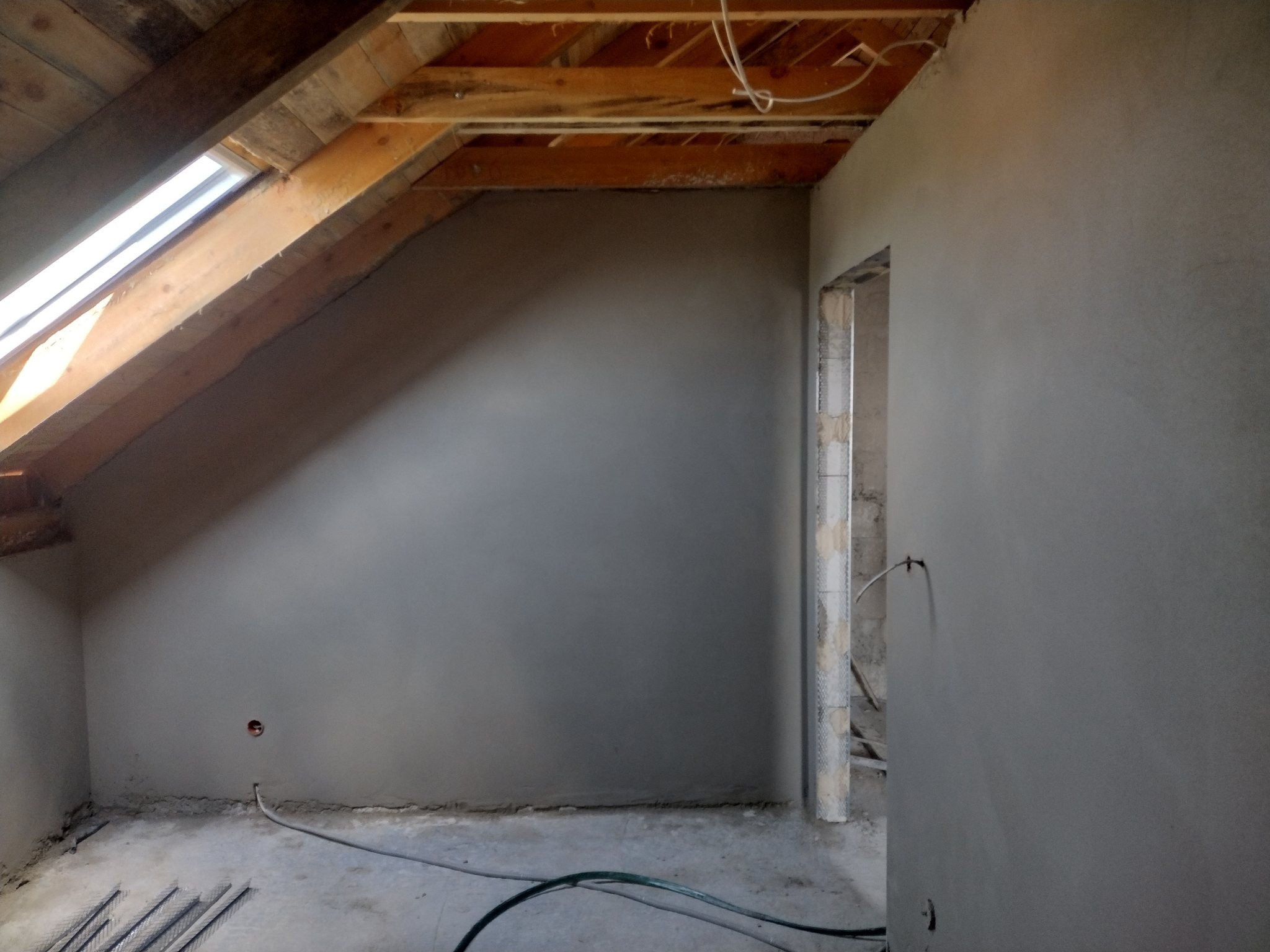 Pokój na poddaszu w trakcie prac wykończeniowych: świeżo położona, gładka warstwa tynku na ścianach, widoczne drewniane belki stropowe, okno dachowe i wystające przewody elektryczne.