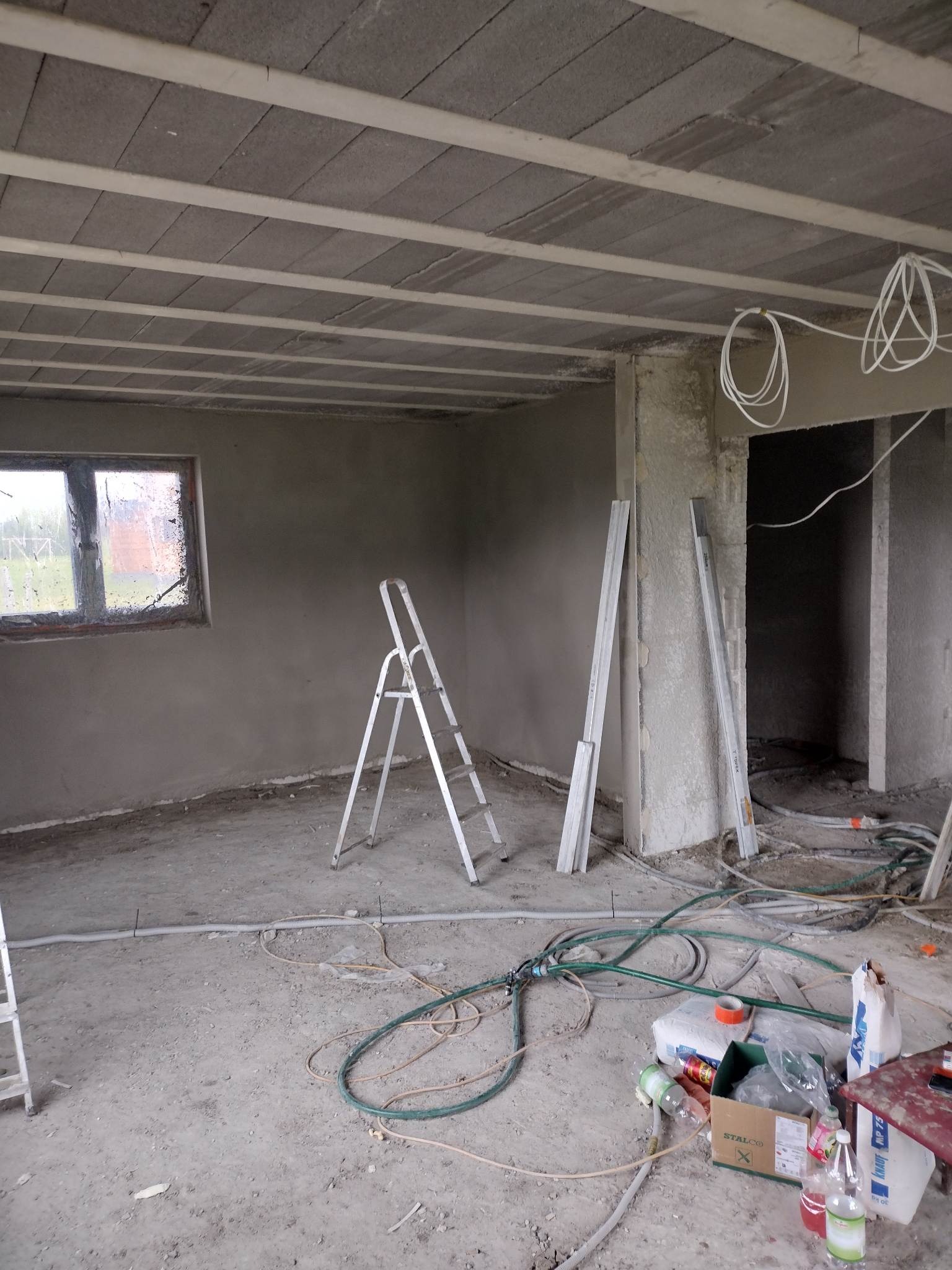 Surowe wnętrze w trakcie prac wykończeniowych: ściany pokryte świeżym tynkiem, aluminiowa drabina, porozrzucane przewody i narzędzia na podłodze, widoczne ślady budowy.