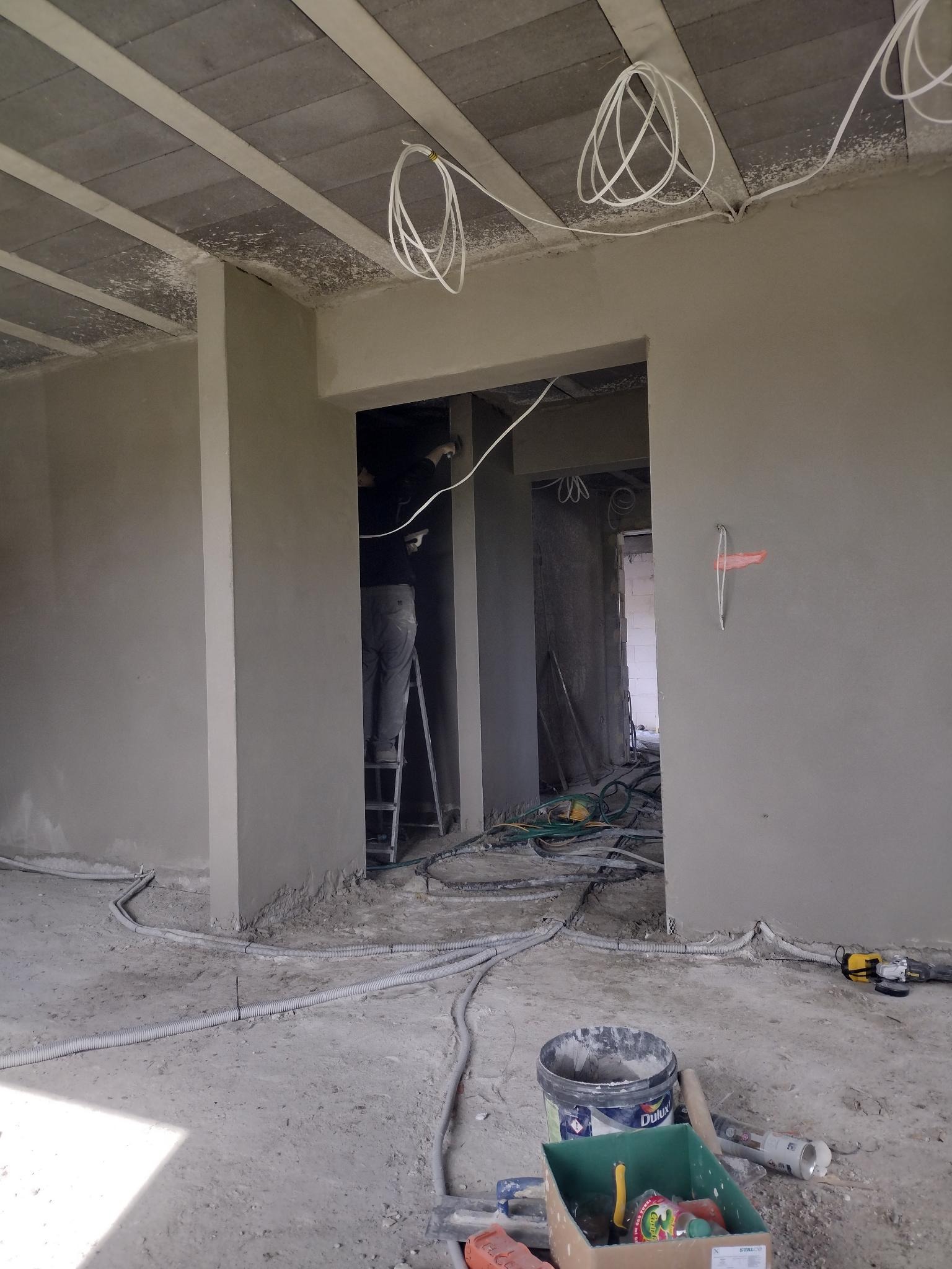 Wnętrze w trakcie prac wykończeniowych: ściany pokryte świeżym tynkiem, widoczne kable elektryczne zwisające z sufitu, pracownik na drabinie w głębi pomieszczenia, na podłodze bałagan i narzędzia.
