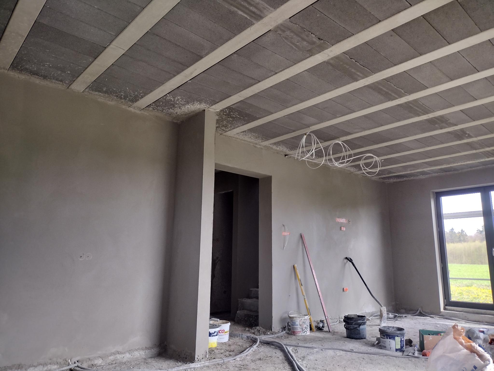Surowe wnętrze w trakcie prac: ściany pokryte szarym tynkiem, widoczne przewody elektryczne zwisające z sufitu, sufit z betonowych płyt, okno z widokiem na zieleń, na podłodze wiadra i narzędzia...