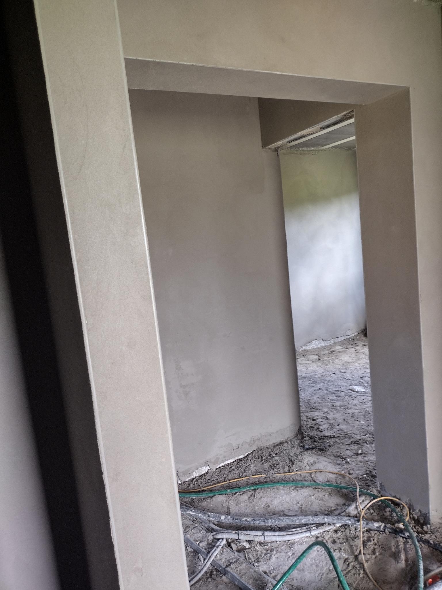 Surowe ściany z nowym tynkiem wokół otworu drzwiowego, widoczne przewody na podłodze w trakcie prac wykończeniowych.