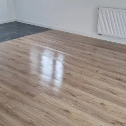 Fragment pokoju z nową podłogą: panele podłogowe o wzorze drewna sąsiadują z ciemnymi, marmurkowymi płytkami przy ścianie, widoczny grzejnik na białej ścianie.