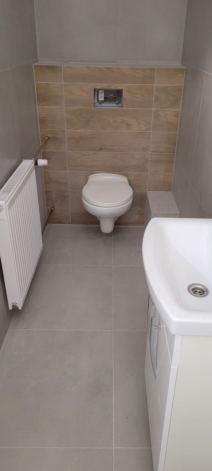 Wykończona łazienka z szarymi płytkami podłogowymi i ściennymi, wisząca toaleta z drewnopodobnymi płytkami za nią, biała umywalka z szafką i grzejnik żeberkowy z miedzianymi rurami.