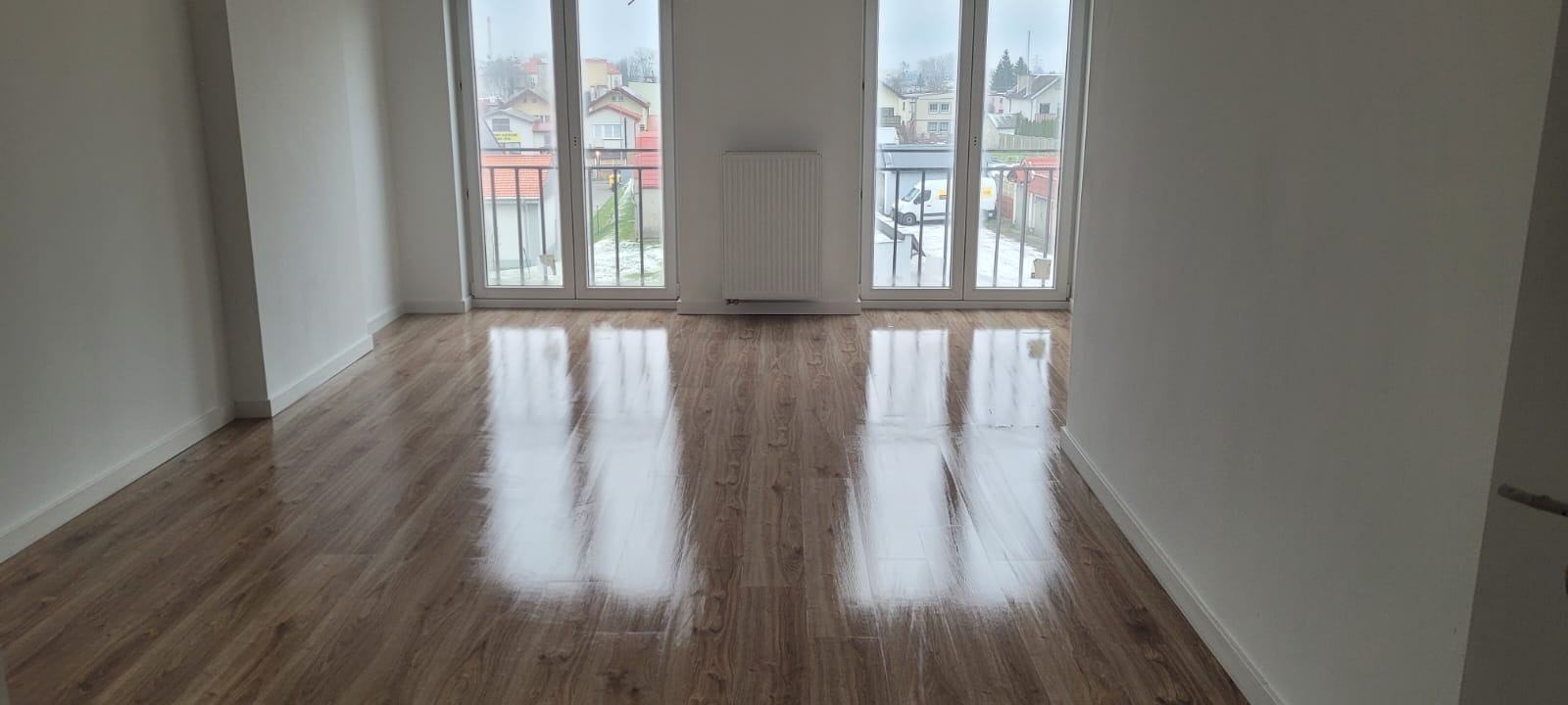 Jasne, puste pomieszczenie z nową podłogą z paneli imitujących drewno i grzejnikiem pod oknem balkonowym, widok na zabudowę jednorodzinną w tle.