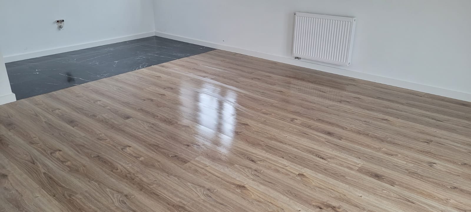 Fragment pokoju z nową podłogą: panele podłogowe o wzorze drewna sąsiadują z ciemnymi, marmurkowymi płytkami przy ścianie, widoczny grzejnik na białej ścianie.