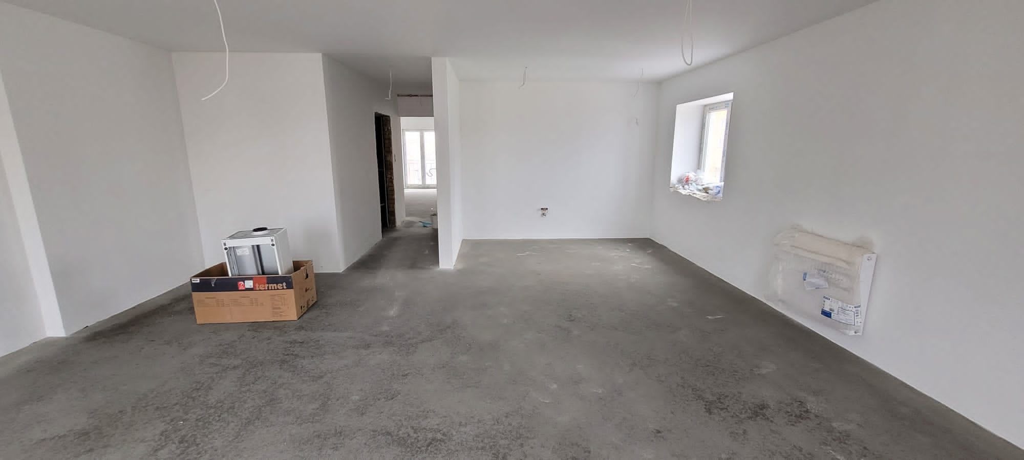Wnętrze w stanie deweloperskim z przygotowaną instalacją grzewczą, widoczne wystające kable, okno z zabezpieczeniem, karton po piecu gazowym Termet i zapakowany grzejnik na ścianie.