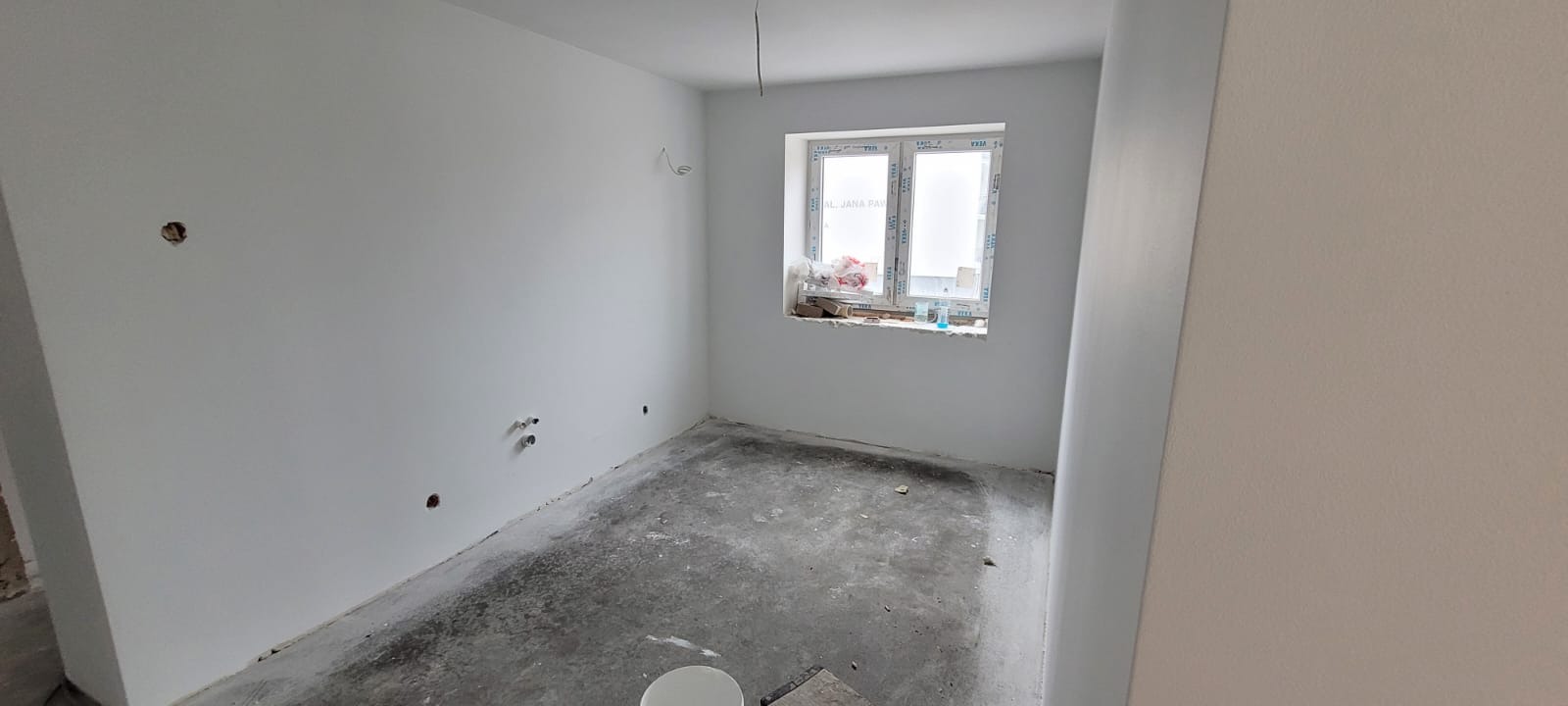 Surowe wnętrze po malowaniu ścian na biało, widoczne wystające rury instalacji hydraulicznej, otwory w ścianach i betonowa wylewka na podłodze.