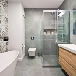 Nowoczesna łazienka z wanną wolnostojącą, kabiną prysznicową ze szklanymi drzwiami przesuwnymi, umywalką z drewnianą szafką i mozaikową ścianą z wnęką.