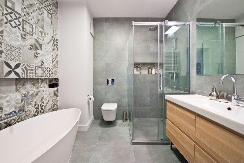 Nowoczesna łazienka z wanną wolnostojącą, kabiną prysznicową ze szklanymi drzwiami przesuwnymi, umywalką z drewnianą szafką i mozaikową ścianą z wnęką.