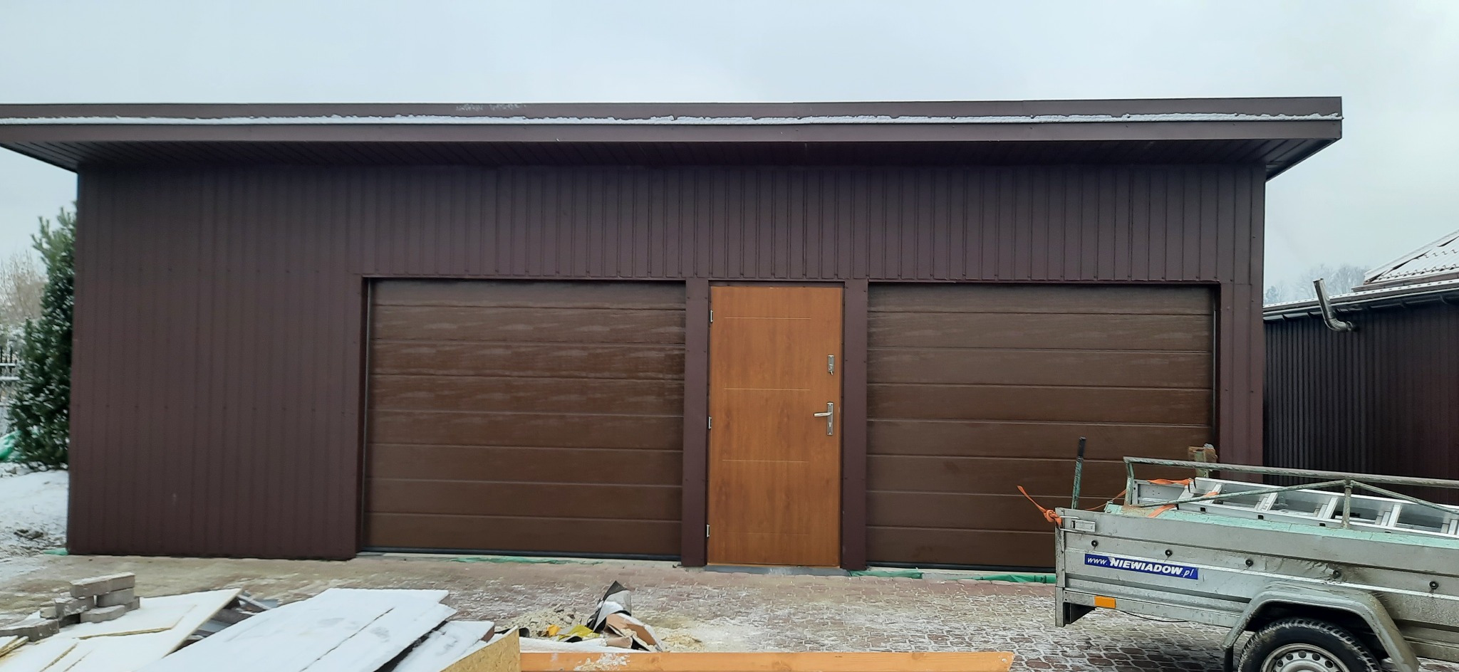 Brązowy budynek gospodarczy z dwiema bramami garażowymi segmentowymi i drzwiami wejściowymi, wykonany z blachy trapezowej, z przyczepką i śniegiem na ziemi.