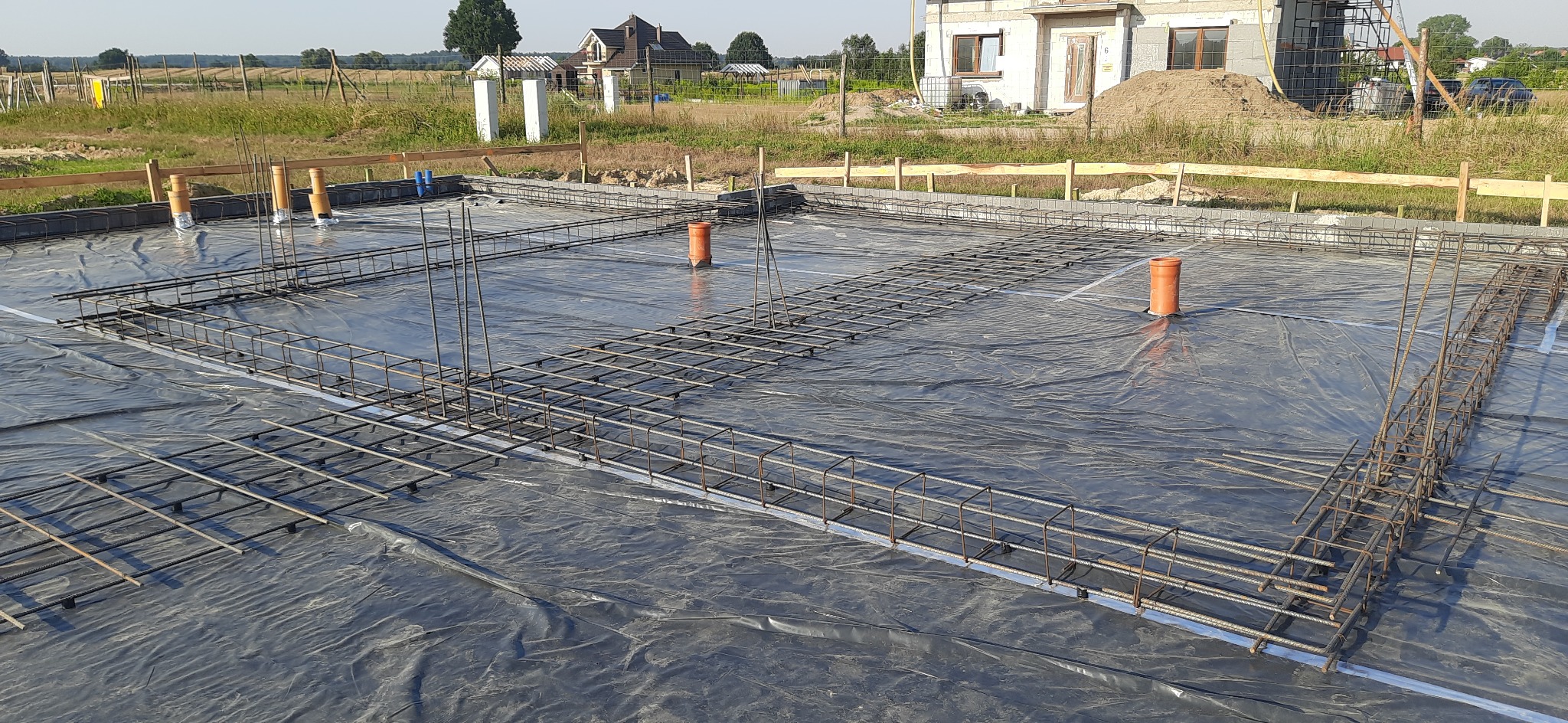 Zbrojenie fundamentów domu jednorodzinnego na folii izolacyjnej, widoczne rury kanalizacyjne, w tle budynek w trakcie budowy.